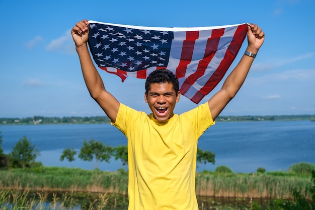 Sonriente joven afroamericano mirando a la cámara y sosteniendo con orgullo la bandera estadounidense ondeando en sus brazos extendidos sobre fondo de cielo azul en verano