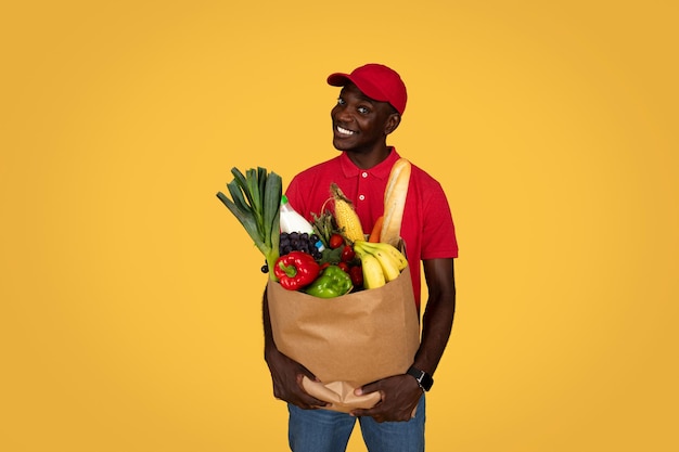 Sonriente joven afroamericano repartidor en uniforme sostiene bolsa ecológica con verduras frutas mira