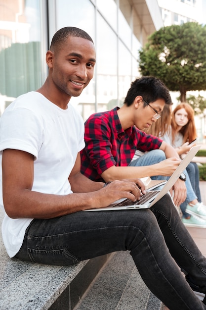 Sonriente joven afroamericano sentado con sus amigos y usando la computadora portátil al aire libre