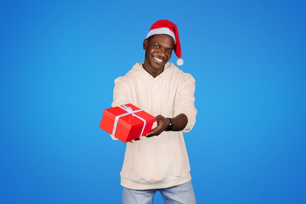 Sonriente joven afroamericano en sudadera Santa gorra sostener caja de regalos aislado en fondo azul