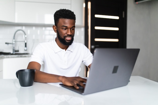Sonriente joven afroamericano tomando café y usando la computadora portátil en la cocina