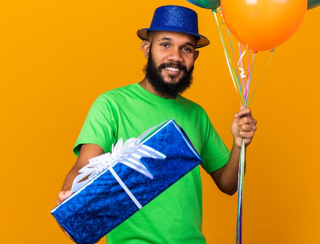 Sonriente joven afroamericano vistiendo gorro de fiesta sosteniendo globos y sosteniendo la caja de regalo en la cámara