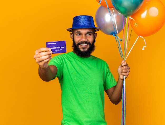 Sonriente joven afroamericano vistiendo gorro de fiesta sosteniendo globos y sosteniendo la tarjeta de crédito en la cámara