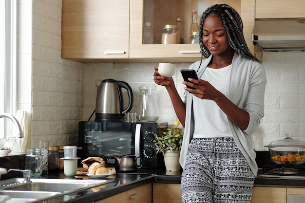 Sonriente joven apoyado en la encimera de la cocina, tomando café y revisando las redes sociales en el teléfono inteligente