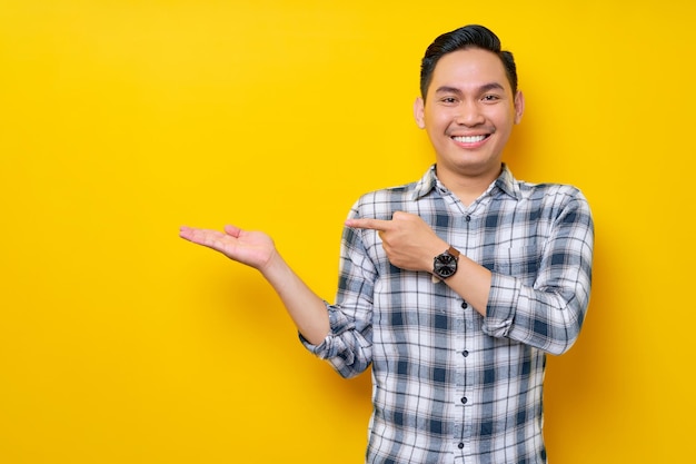 Sonriente joven apuesto hombre asiático de 20 años con una camisa a cuadros apuntando con el dedo a la palma abierta anunciando algo aislado en el fondo amarillo Concepto de estilo de vida de la gente