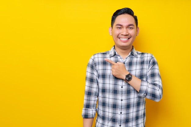 Sonriente joven apuesto hombre asiático de 20 años con una camisa a cuadros señalando con el dedo el área de publicidad aislada sobre fondo amarillo Concepto de estilo de vida de la gente