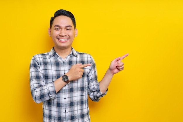 Sonriente joven apuesto hombre asiático de 20 años con una camisa a cuadros señalando con el dedo el área de publicidad aislada sobre fondo amarillo Concepto de estilo de vida de la gente