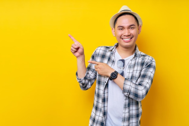 Sonriente joven apuesto hombre asiático étnico de 20 años con ropa informal sombrero de pie señalando con el dedo a un lado en el espacio de copia simulada aislado en fondo amarillo Concepto de estilo de vida de la gente