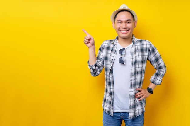 Sonriente joven apuesto hombre asiático étnico de 20 años con ropa informal sombrero de pie señalando con el dedo a un lado en el espacio de copia simulada aislado en fondo amarillo Concepto de estilo de vida de la gente