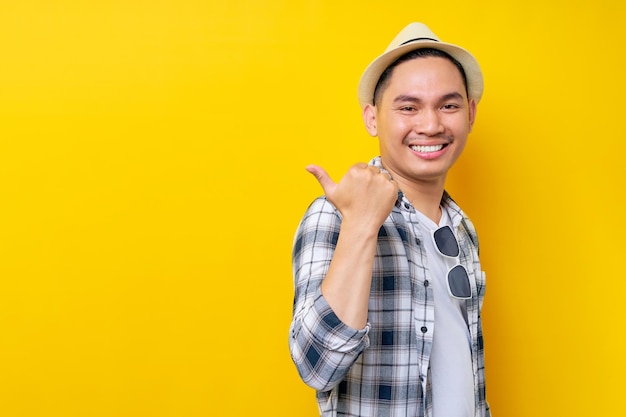 Sonriente joven apuesto hombre asiático étnico de 20 años con ropa informal sombrero de pie señalando con el pulgar a un lado en el espacio de copia simulada aislado en fondo amarillo Concepto de estilo de vida de la gente