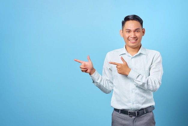 Sonriente joven apuesto hombre de negocios apuntando con el dedo hacia el espacio de la copia sobre fondo azul.