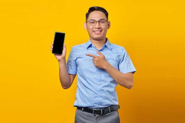 Sonriente joven apuesto hombre de negocios en camisa azul clásica apuntando con el dedo al teléfono inteligente de pantalla en blanco aislado sobre fondo amarillo Lograr el concepto de negocio de riqueza profesional