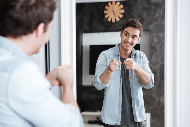 Sonriente joven apuntando con el dedo a su reflejo en el espejo mientras está de pie en casa