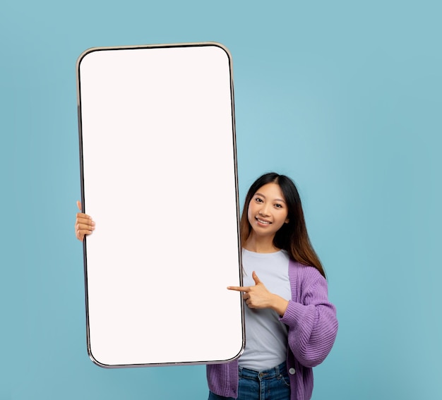 Sonriente joven asiática apuntando a un gran celular con pantalla vacía sobre el fondo azul del estudio