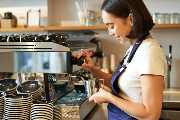 Sonriente joven asiática barista chica usando máquina de café vapor leche humeante para capuchino orden sta