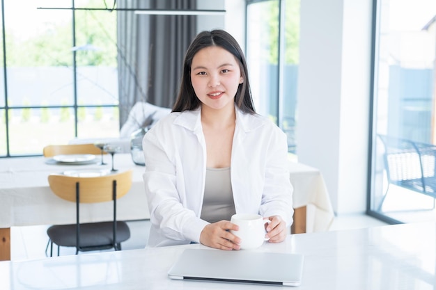 sonriente joven asiática con camisa blanca con taza de té o café y portátil en casa moderna