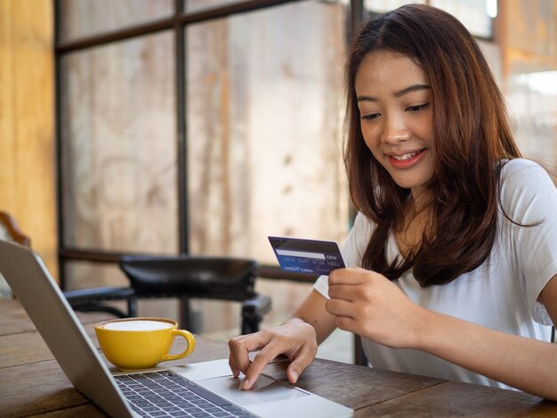 La sonriente joven asiática disfruta comprando en línea a través de una computadora y pagando en línea con tarjeta de crédito. Conveniencia de gastar sin efectivo. manténgase seguro, comprando desde casa y a distancia social