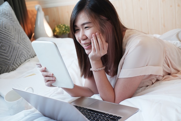 Sonriente joven asiática está acostado en la cama blanca con tableta para trabajar en línea y hacer compras con el portátil en la medea social en el dormitorio. La empresaria trabaja desde casa con el concepto de tecnología.