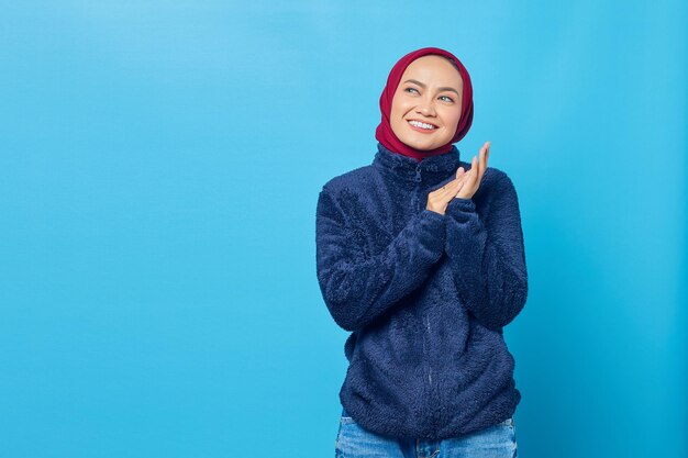 Sonriente joven asiática frotar las manos y mirando a un lado sobre fondo azul.