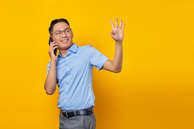 Sonriente joven asiático asiático con anteojos hablando por teléfono móvil mientras agita el gesto de la mano aislado en el concepto de empresario y empresario de fondo amarillo