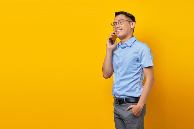 Sonriente joven asiático asiático con anteojos hablando por teléfono móvil y mirando a un lado aislado en el concepto de empresario y empresario de fondo amarillo