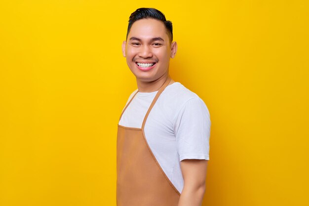 Sonriente joven asiático barista barman empleado de 20 años con delantal marrón trabajando en una cafetería con confianza en la cámara aislada en fondo amarillo Concepto de inicio de pequeñas empresas