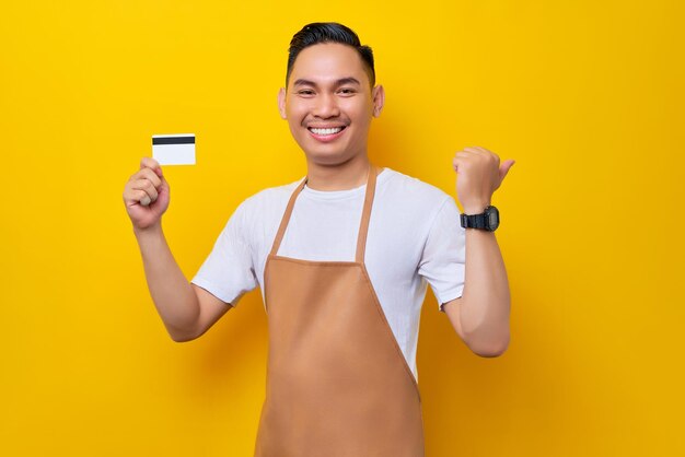 Sonriente joven asiático barista barman empleado de 20 años con delantal marrón trabajando en una cafetería con tarjeta de crédito apuntando el pulgar a un lado sobre fondo amarillo Concepto de inicio de pequeñas empresas