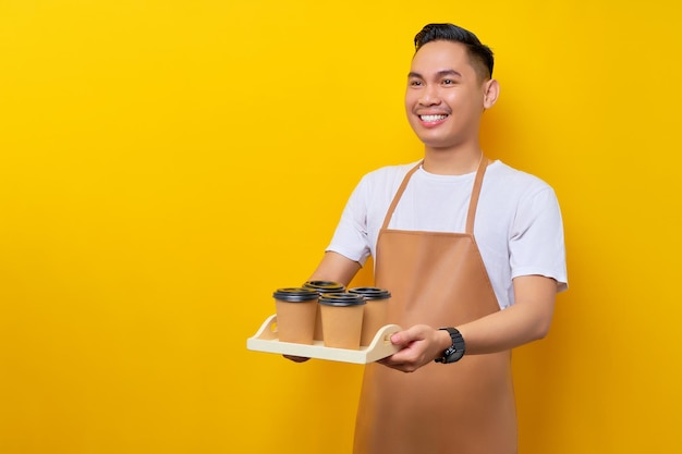 Sonriente joven asiático barista barman empleado con delantal marrón trabajando en una cafetería dando tazas de café o té para llevar y mirando a un lado sobre fondo amarillo Pequeña empresa nueva