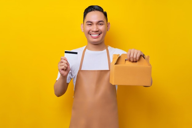 Sonriente joven asiático barista barman empleado con delantal marrón trabajando en una cafetería sosteniendo una bolsa de pastel de papel para llevar y una tarjeta de crédito con fondo amarillo Concepto de inicio de una pequeña empresa
