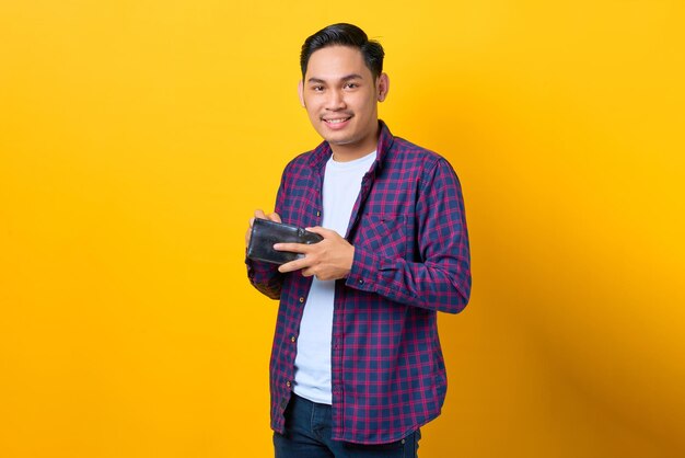 Sonriente joven asiático con camisa a cuadros poniendo dinero en su billetera aislado de fondo amarillo