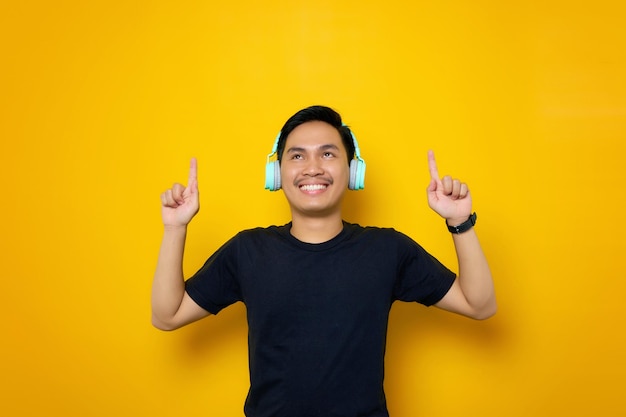 Sonriente joven asiático en camiseta casual con auriculares apuntando con el dedo al espacio de copia aislado sobre fondo amarillo Gente emociones concepto de estilo de vida