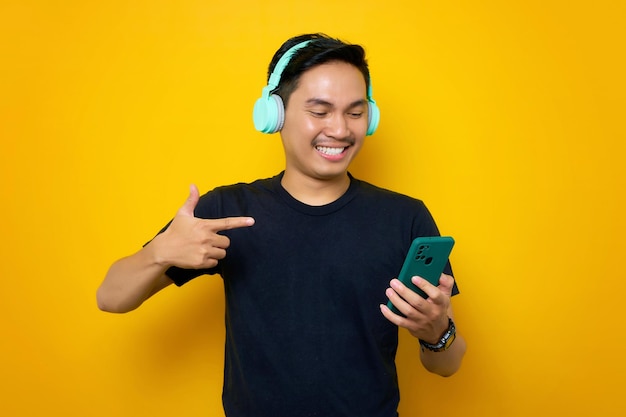 Sonriente joven asiático en camiseta casual con auriculares inalámbricos apuntando con el dedo al teléfono móvil aislado sobre fondo amarillo Gente emociones concepto de estilo de vida