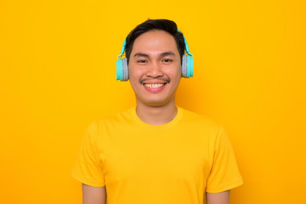 Sonriente joven asiático en camiseta casual escuchando música en auriculares mirando a la cámara aislada sobre fondo amarillo Personas emociones concepto de estilo de vida