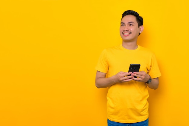 Sonriente joven asiático en camiseta casual sosteniendo teléfono móvil y mirando a un lado aislado sobre fondo amarillo Concepto de estilo de vida de la gente