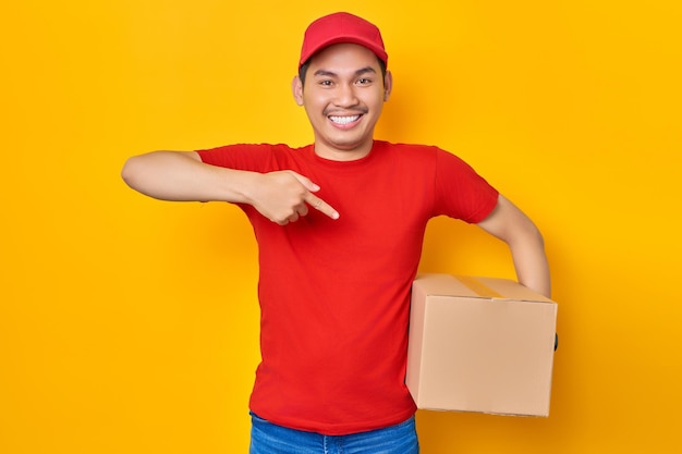 Sonriente joven asiático con gorra roja, camiseta, uniforme, empleado, trabajo como distribuidor, mensajero, llevando, paquete, caja, recomendación, tienda, aislado, en, fondo amarillo, profesional, entrega, servicio, concepto