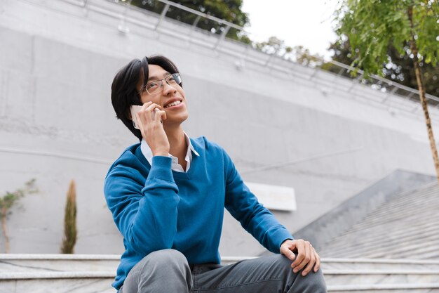 Sonriente joven asiático hablando por teléfono móvil mientras está sentado en la escalera al aire libre