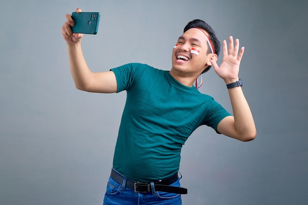 Sonriente joven asiático haciendo selfie tiro en teléfono móvil agitando la mano aislado sobre fondo gris Concepto de celebración del día de la independencia de Indonesia