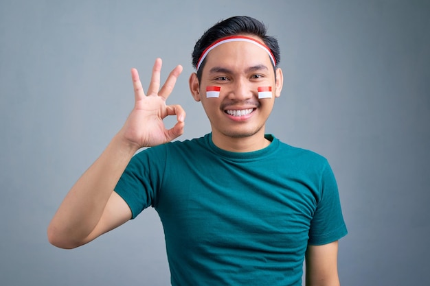 Sonriente joven asiático mostrando ok firmar celebrando el día de la independencia de Indonesia aislado sobre fondo gris Concepto de celebración del día de la independencia de Indonesia