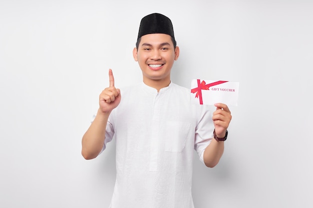 Sonriente joven asiático musulmán sosteniendo un vale de certificado de regalo apuntando con su dedo hacia arriba aislado en fondo blanco Gente religiosa Islam estilo de vida concepto celebración Ramadán e ie Mubarak