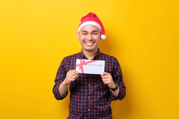 Sonriente joven asiático con sombrero de Papá Noel sosteniendo un certificado de regalo mirando a la cámara sobre el fondo amarillo del estudio Feliz Año Nuevo 2023 celebración feliz concepto de vacaciones