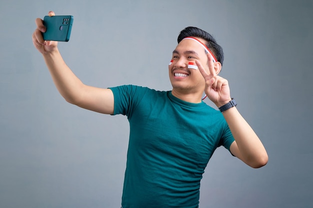 Sonriente joven asiático tomando selfie en el teléfono móvil con hacer el signo de la paz aislado sobre fondo gris Concepto de celebración del día de la independencia de Indonesia