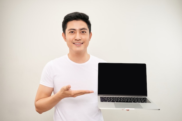 Sonriente joven asiático vestido con camisa mostrando ordenador portátil con pantalla en blanco sobre fondo blanco.
