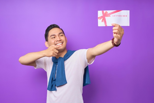 Sonriente joven asiático vestido con ropa informal señalando con el dedo el certificado de vales de regalo y mirando la cámara aislada en el fondo del estudio morado