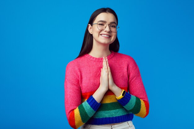 Sonriente joven atractiva mostrar gesto de oración viste un suéter de arco iris