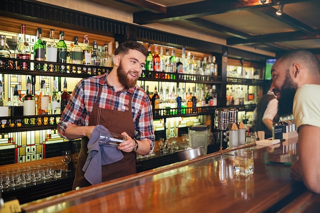 Sonriente joven atractivo barman limpiando vasos y hablando con el joven en el bar