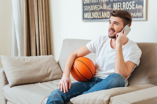 Sonriente joven atractivo sosteniendo una pelota de baloncesto y hablando por teléfono celular en casa