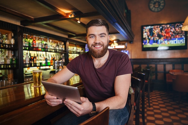 Sonriente joven atractivo con tableta y bebiendo cerveza en el pub