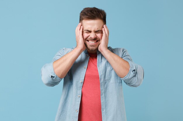 Sonriente joven barbudo de 20 años con camisa informal posando aislado en un retrato de estudio de fondo azul pastel. Concepto de estilo de vida de las emociones de las personas. Simulacros de espacio de copia. Manteniendo los ojos cerrados ponga las manos en las mejillas.