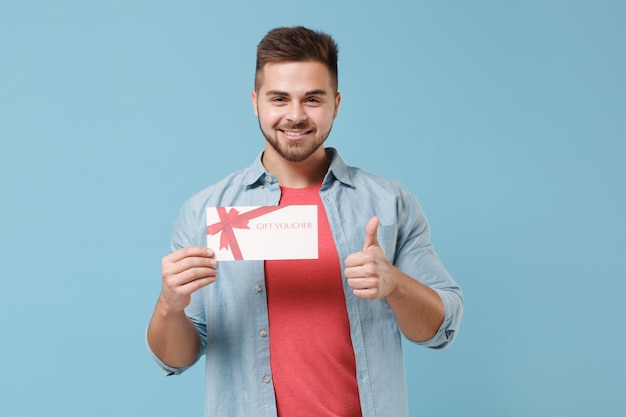 Sonriente joven barbudo de 20 años con camisa informal posando aislado en un retrato de estudio de fondo azul pastel. Concepto de estilo de vida de las emociones de las personas. Simulacros de espacio de copia. Sostenga el certificado de regalo, mostrando el pulgar hacia arriba.