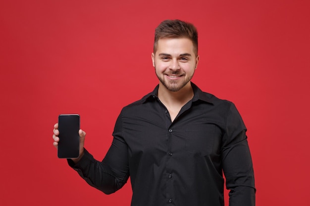 Sonriente joven barbudo de 20 años con camisa negra clásica posando aislado en un retrato de estudio de fondo de pared roja. Concepto de estilo de vida de las personas. Simulacros de espacio de copia. Sostenga el teléfono móvil con la pantalla vacía en blanco.